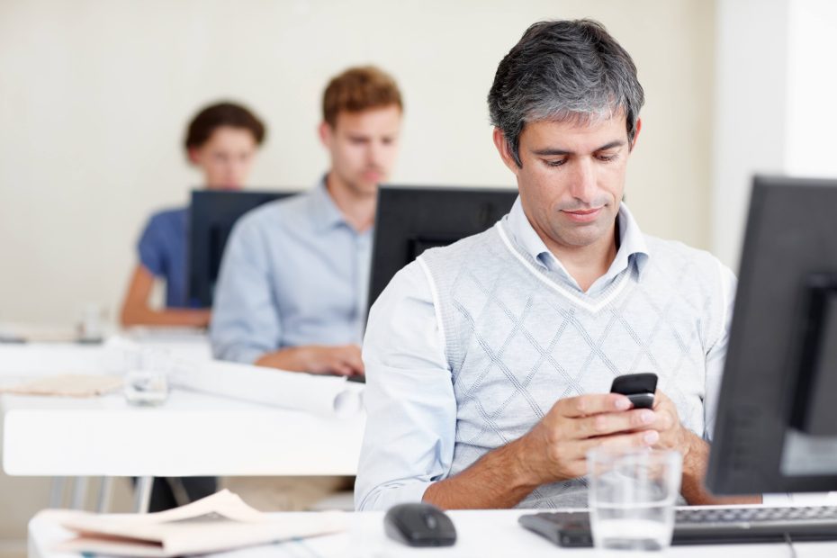 Row of three men working on computers, the man in front is looking at his smart phone